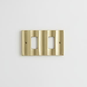 Pode incluir: Uma placa de interruptor dupla de cor latão com um design ondulado e texturizado. A placa tem duas aberturas retangulares para os interruptores e é montada em uma parede branca. A placa é fixada com parafusos.