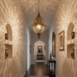 May include: A decorative gold pendant light hangs in a hallway, creating detailed shadow patterns on the walls. The hallway has a patterned rug, arched wall niches with vases, and dark wood flooring. The overall design is warm and welcoming.