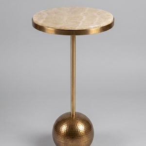 May include: A side table featuring a circular top made of light beige, translucent stone with natural veining. The table is supported by a slender, vertical brass-toned metal rod connected to a heavy, spherical base with a hammered texture.