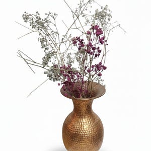 May include: A hammered copper vase with a wide, flared rim holds a bouquet of dried flowers. The flowers include sprigs of white and purple blooms. The vase has a textured surface and a round body.