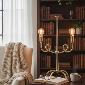 May include: A brass candelabra-style lamp with two Edison-style bulbs illuminates a cozy reading nook. The lamp sits on a dark wood table with an open book and eyeglasses. A beige knitted throw covers an armchair, and a bookcase filled with books is in the background.