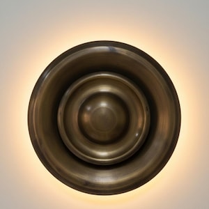 May include: A bronze-colored wall sconce with a circular design. The sconce has a layered, bowl-like shape with a central, rounded element. The warm light emanating from behind the sconce creates a halo effect, highlighting its metallic finish.