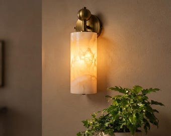 May include: A wall-mounted light fixture featuring a cylindrical shade made of translucent, cream-colored stone with natural veining. The fixture is attached to a circular brass-toned metal base with a spherical accent piece at the top.