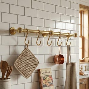 May include: A kitchen scene featuring a brass hanging rack with a pot holder, copper measuring cup, and striped apron. A cookbook and red and white checked tea towel sit on a wooden shelf. Wooden utensils are in a ceramic pot.