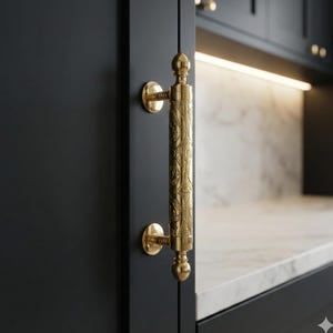 Könnte beinhalten: Nahaufnahme eines goldfarbenen Schrankgriffs mit einem verzierten, gravierten Design. Der Griff ist an einer dunkelgrauen Schranktür befestigt. Die Hardware hat einen klassischen, eleganten Stil mit einem zylindrischen Griff und dekorativen Endstücken.
