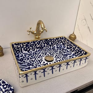 Puede incluir: Lavabo rectangular de cerámica con interior estampado en azul y blanco y borde dorado. El lavabo tiene un grifo dorado y una tapa de desagüe decorativa. Es un accesorio decorativo para el baño.