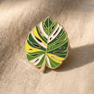 May include: A colorful enamel pin in the shape of a monstera leaf. The pin features a gold outline with sections of green, yellow, white, and black. The pin is a botanical accessory.