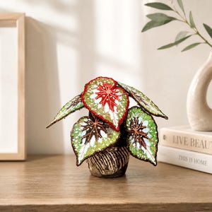 Peut inclure: Une sculpture de plante décorative présentant des feuilles en forme de cœur avec des motifs perlés complexes rouges, blancs et verts. La plante est placée dans un petit pot brun foncé texturé avec une finition rayée, montrant un travail détaillé.