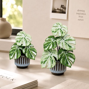 May include: Two decorative potted plants featuring embroidered monstera leaves in shades of green and white. Each plant is set in a small, vertically ribbed ceramic pot with a light blue glaze at the base and a dark brown finish at the top rim.