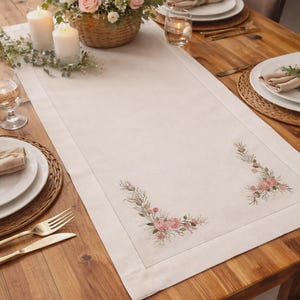 May include: A rectangular off-white table runner featuring delicate embroidered floral motifs in shades of pink and green at both ends. The fabric has a visible woven texture with a stitched border detail, laid flat on a wooden dining table.