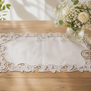 May include: A white linen table runner with intricate lace detailing along the edges. The runner is placed on a wooden table, with a vase of white flowers and a candle holder in the background. The runner's dimensions are approximately 12 inches wide and 24 inches long.