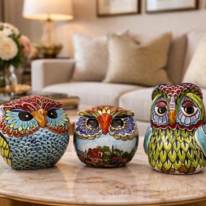 Peut inclure: Trois figurines de hiboux colorés avec des détails peints complexes. Les hiboux sont exposés sur une table en marbre devant un canapé aux tons neutres. Chaque hibou présente un design unique, avec des bleus, verts et rouges vibrants.