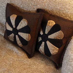 May include: Two brown decorative cushions with a floral design. Each cushion features a black centre and alternating tan and black petals. The cushions are placed on a textured, light brown surface.