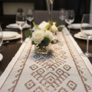 May include: A dining table set for a meal, featuring a cream-colored table runner with a geometric pattern. White roses in a glass vase and wine glasses are arranged on the dark wood table. White plates are also set.