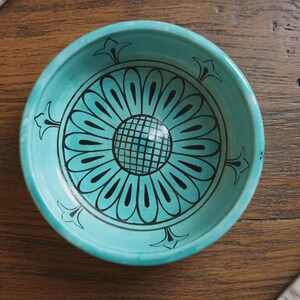 May include: A round turquoise ceramic bowl featuring a black hand-painted floral pattern on the interior. The design consists of a central grid-patterned circle surrounded by elongated petals and four small decorative flourishes along the rim.