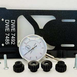 Könnte beinhalten: Eine schwarze Metallhalterung mit dem Text "DWE 7492 DWE 7485" und eine Messuhr mit weißem Zifferblatt. Enthält auch mehrere schwarze Knöpfe und silberne Schrauben, wahrscheinlich für Präzisionsmessungen oder Kalibrierungen.