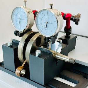 May include: A precision measuring tool featuring two dial indicators, mounted on a black base. The gauges have white faces with black markings and measure in 0.01mm increments. The setup includes a crankshaft and connecting rod, used for engine component alignment.