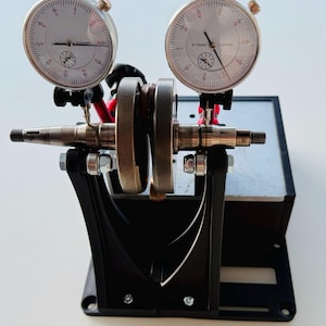 Può includere: Uno strumento di allineamento per albero motore in metallo con due indicatori a quadrante circolari con scale di misura 0-10mm. Il dispositivo include una base nera, due supporti verticali e bracci rossi regolabili che tengono i calibri.