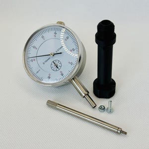 Könnte beinhalten: Ein Messuhr mit weißem Zifferblatt, schwarzen Markierungen und einem silbernen Metallgehäuse. Das Zifferblatt zeigt 0-10mm und 0,01mm an. Ein schwarzes zylindrisches Teil, eine silberne Stange und kleine Hardware sind ebenfalls vorhanden.