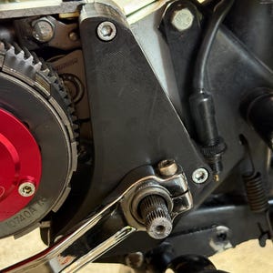 May include: Close-up of motorcycle engine components. The image shows a black bracket, silver bolts, and a red and silver gear assembly. A chrome wrench is also visible, along with various wires and tubes.