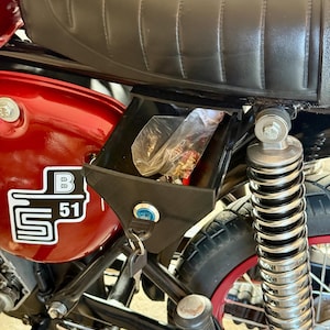 Könnte beinhalten: Nahaufnahme eines roten Motorrads mit schwarzem Sitz und Staufach. Das Staufach befindet sich unter dem Sitz und hat ein Schloss mit einem Schlüssel. Das Motorrad hat ein "B 51" Logo an der Seite.