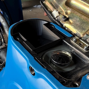 Op de afbeelding: Close-up van een blauwe scooter carrosserie met een zwart compartiment en een chromen tankdop. De zitting van de scooter is open, waardoor de interne componenten zichtbaar zijn. De afbeelding benadrukt het ontwerp en de mechanische elementen van de scooter.