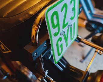May include: Close-up of a motorcycle's rear section, showcasing a black seat with a quilted pattern. A chrome bar supports a green and white license plate with the numbers "923 GYU". The plate is mounted on a wooden bracket.