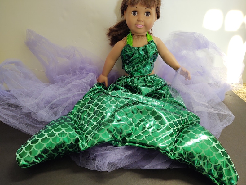Puede incluir: Un disfraz de cola de sirena verde para una mu&ntilde;eca. La cola est&aacute; hecha de tela brillante con un patr&oacute;n de escamas. La mu&ntilde;eca lleva un top verde con un collar verde y blanco.