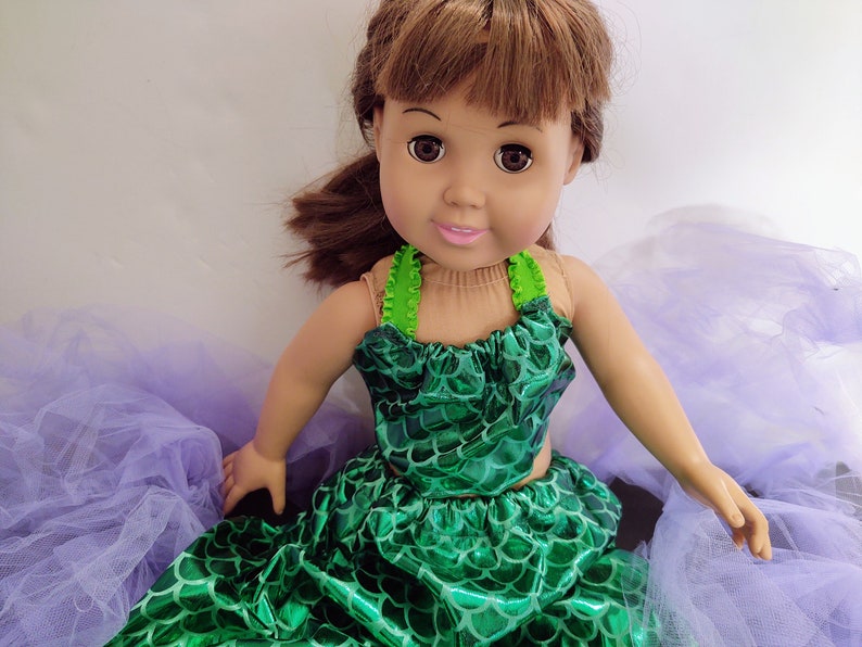 Puede incluir: Una mu&ntilde;eca con un disfraz de sirena verde y una falda de tul morado. La mu&ntilde;eca tiene el pelo casta&ntilde;o y los ojos marrones.