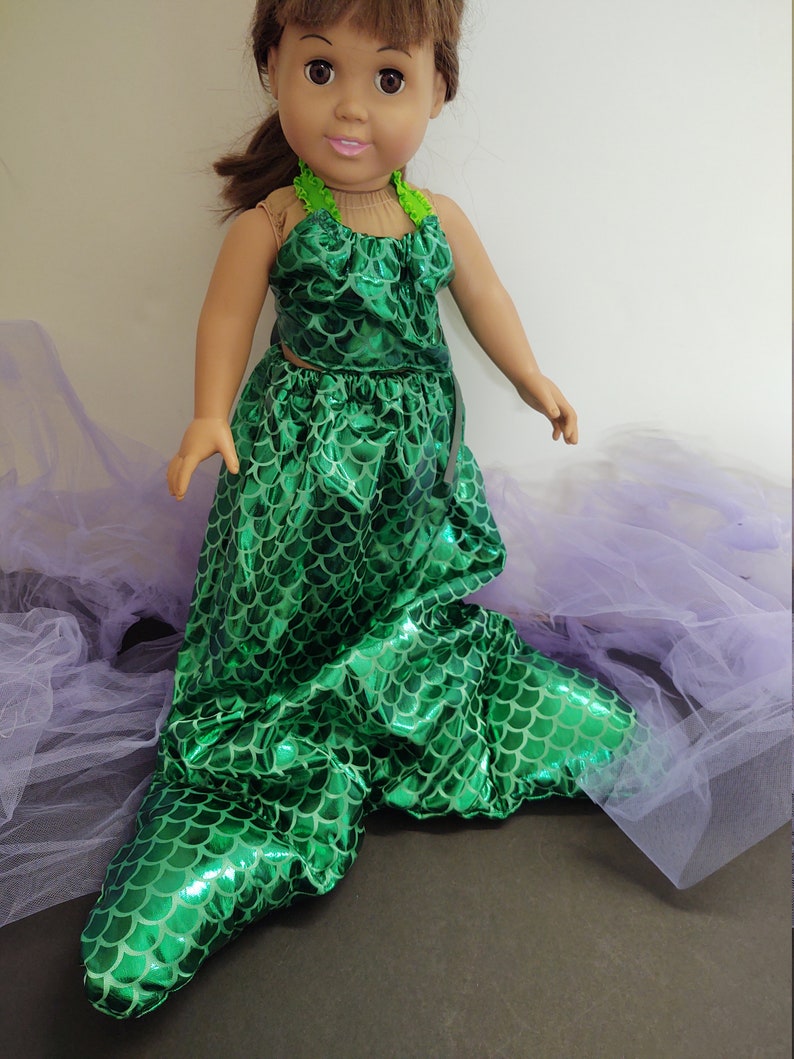 Puede incluir: Un disfraz de sirena verde para una mu&ntilde;eca. El disfraz presenta un top y una cola de color verde iridiscente con estampado de escamas. La cola es larga y fluida, con una aleta puntiaguda en el extremo.