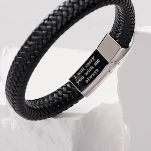 May include: Black braided leather bracelet with a silver clasp. The clasp is engraved with the words "I will carry you with me always". The bracelet is displayed on a white surface.