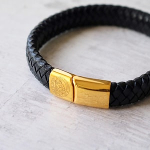 May include: A black braided leather bracelet featuring a polished gold-tone metal clasp. The clasp is engraved with a circular Saint Christopher design and the text "Saint Christopher Protect Us" on one side, providing a secure closure.