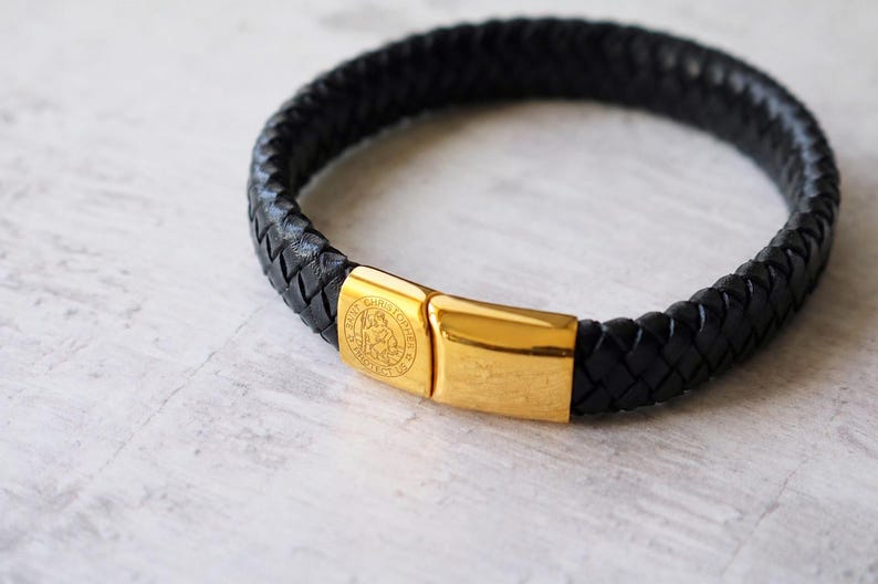 May include: A black braided leather bracelet featuring a polished gold-tone metal clasp. The clasp is engraved with a circular Saint Christopher design and the text "Saint Christopher Protect Us" on one side, providing a secure closure.