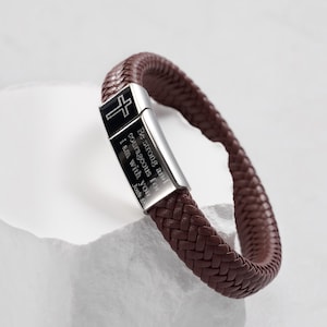 May include: A brown braided leather bracelet featuring a rectangular silver-tone metal clasp. The clasp is engraved with a black cross symbol and the text: Be strong and courageous for i am with you Josh 1:9.
