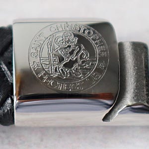 May include: A black braided bracelet featuring a polished silver-tone metal clasp. The clasp is engraved with a circular design depicting Saint Christopher carrying a child, surrounded by the text SAINT CHRISTOPHER PROTECT US.