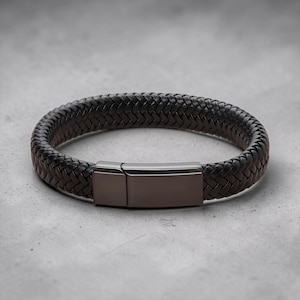 May include: A dark brown braided leather bracelet featuring a smooth, rectangular gunmetal-toned metal clasp. The bracelet has a thick, woven texture and a polished finish on the metallic closure, creating a clean and simple design.
