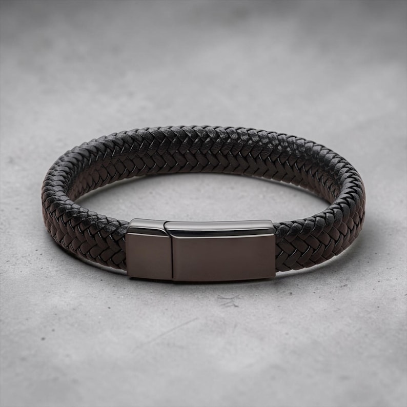 May include: A dark brown braided leather bracelet featuring a smooth, rectangular gunmetal-toned metal clasp. The bracelet has a thick, woven texture and a polished finish on the metallic closure, creating a clean and simple design.