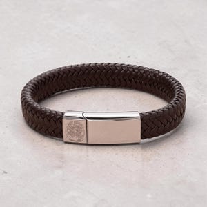 May include: A dark brown braided leather bracelet featuring a polished silver-tone metal clasp. The clasp is rectangular with a smooth surface and includes a small circular engraving of Saint Michael on the left side of the metal closure.