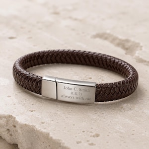 Puede incluir: Una pulsera de cuero trenzado marrón oscuro con un cierre rectangular de metal plateado. El cierre está grabado con el texto John C. Smith, 11.11.21, y always with me. La pulsera tiene una textura tejida y un acabado metálico pulido.