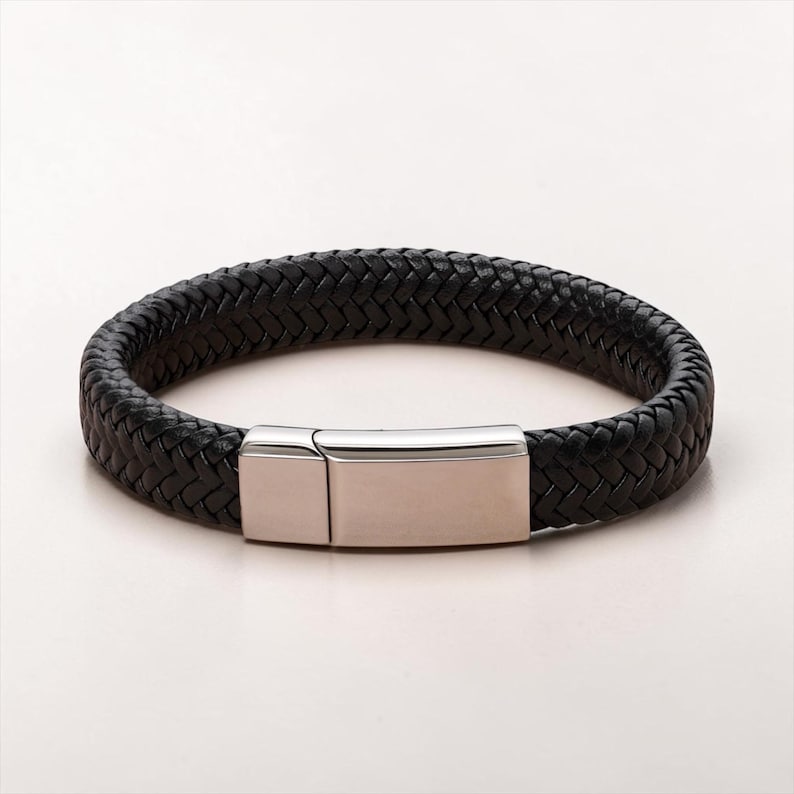 May include: A black braided leather bracelet featuring a polished silver-tone metal rectangular clasp. The bracelet has a thick, woven texture and a smooth, metallic closure that secures the two ends of the leather band together in a loop.
