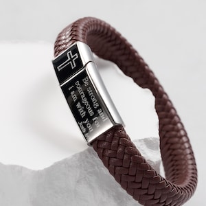 May include: A brown leather bracelet with a woven design. The silver clasp features a black cross and the engraved text "Be strong and courageous for I am with you Josh 1:9". A stylish accessory.