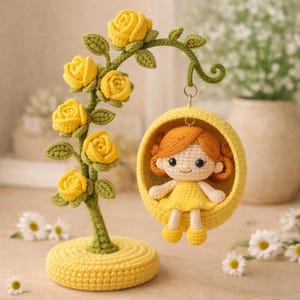 May include: A handcrafted crochet decorative piece featuring a yellow rose tree and a hanging yellow pod with a doll inside. The doll has orange hair and a yellow dress. The base of the tree is yellow, and the leaves are green.