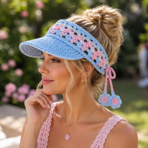 Könnte beinhalten: Ein hellblauer Häkel-Sonnenblende mit einem pastellrosa und weißen Musterband. Die Sonnenblende hat einen einfarbig blauen Schirm und zwei baumelnde Häkelblumen-Anhänger in Rosa und Blau, die mit dünnen rosa Bändern an der Seite befestigt sind.
