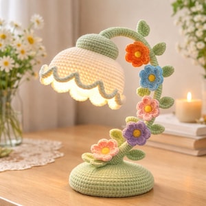 May include: A handcrafted crochet table lamp in a floral design. The lamp features a light green base and stem, with a white scalloped lampshade. Colorful crochet flowers in orange, blue, pink, and purple adorn the stem, adding a whimsical touch.