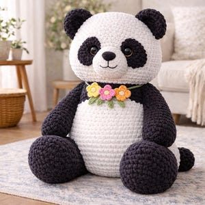 May include: A handmade crochet panda bear with black and white yarn. The panda has black eyes, ears, and limbs, with a white body and head. A floral necklace of yellow, pink, and orange flowers adorns its neck. The panda is sitting on a patterned rug.