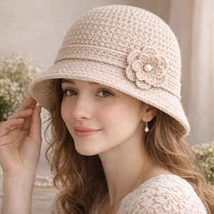 Puede incluir: Sombrero cloche de ganchillo beige con una flor decorativa y una perla en el centro. El sombrero tiene un ala y una banda alrededor de la corona. El sombrero es de estilo vintage y es adecuado para diversas ocasiones.