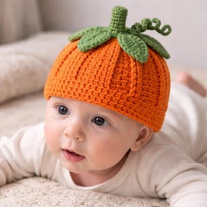 May include: An orange crochet pumpkin hat with green leaves and stem. The hat is worn by a baby lying on a soft, textured surface. The baby is wearing a white long-sleeved shirt.
