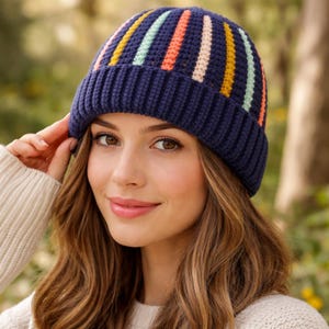 May include: A navy blue knit beanie with vertical stripes in yellow, light blue, orange, pink, and white. The hat has a ribbed brim and is worn by a person with long brown hair.