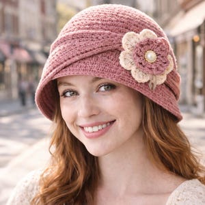 Könnte beinhalten: Ein staubrosa gehäkelter Glockenhut mit einer dekorativen Blume. Der Hut hat eine breite Krempe und einen floralen Akzent mit einem Perlenmittelpunkt. Ein stilvolles Accessoire für jedes Outfit.