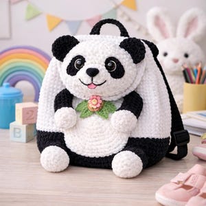 May include: A white and black crochet panda bear backpack with a black handle and straps. The panda has black ears, eyes, and paws, with a pink flower and green leaves on its chest. The backpack is designed for children.